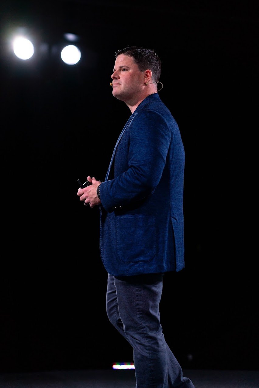 Jordan Shaw speaking on stage in a blue blazer, profile lit against darkness
